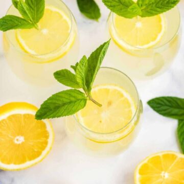 Three glasses of lemonade garnished with lemon slices and mint leaves on a marble surface, with whole and sliced lemons nearby.