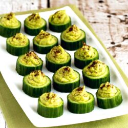 Slices of cucumber topped with a dollop of green avocado mixture, garnished with spices, arranged neatly on a rectangular white plate.