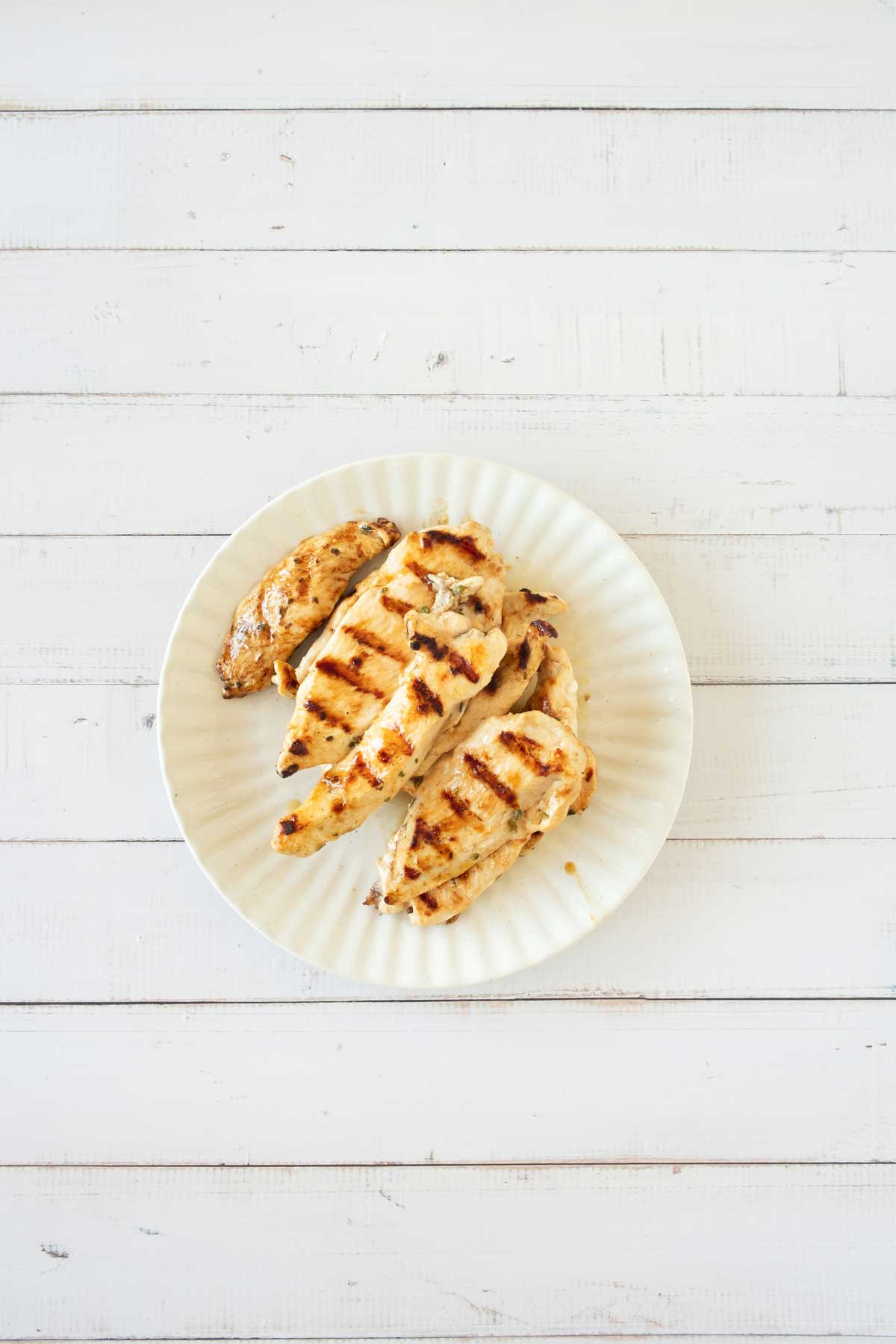 Grilled chicken breasts with grill marks on a white plate, placed on a white wooden surface.