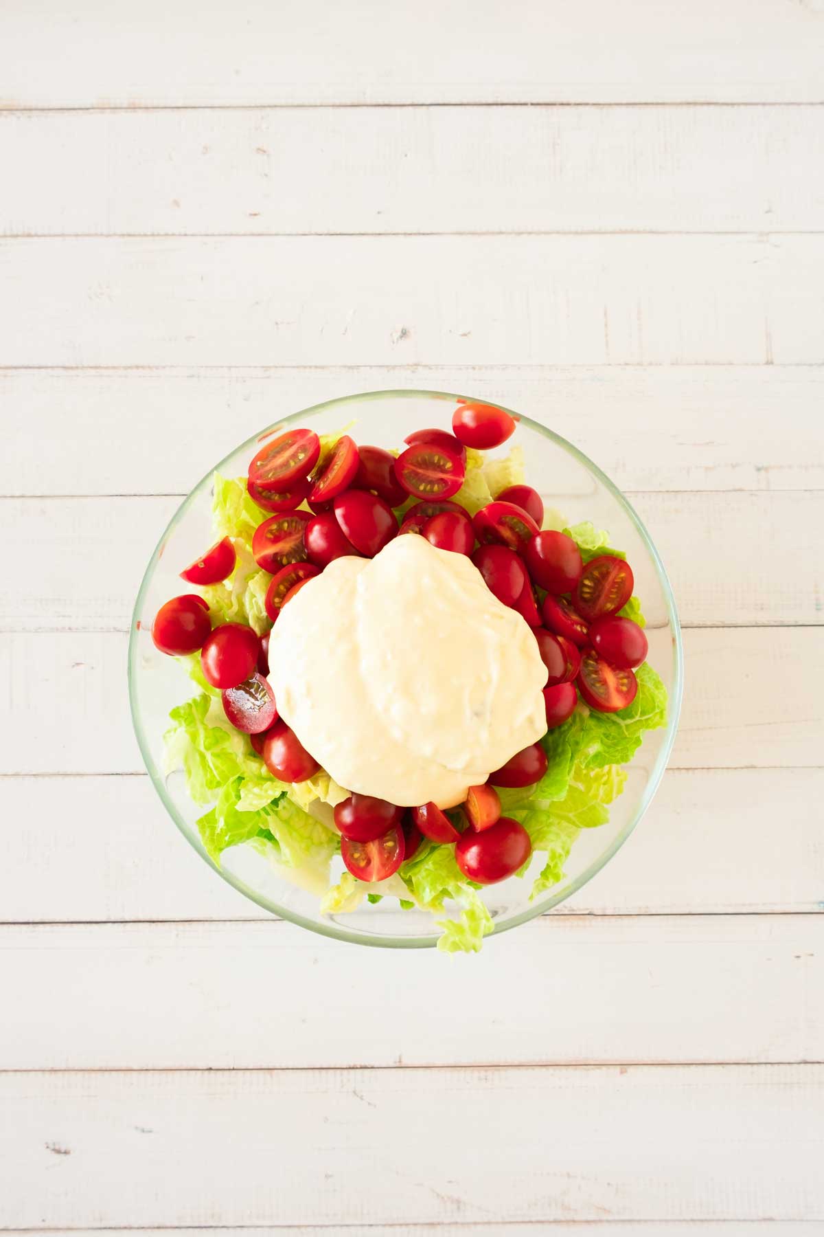 A bowl of salad with lettuce, halved cherry tomatoes, and a scoop of mayonnaise on top, placed on a white wooden surface.