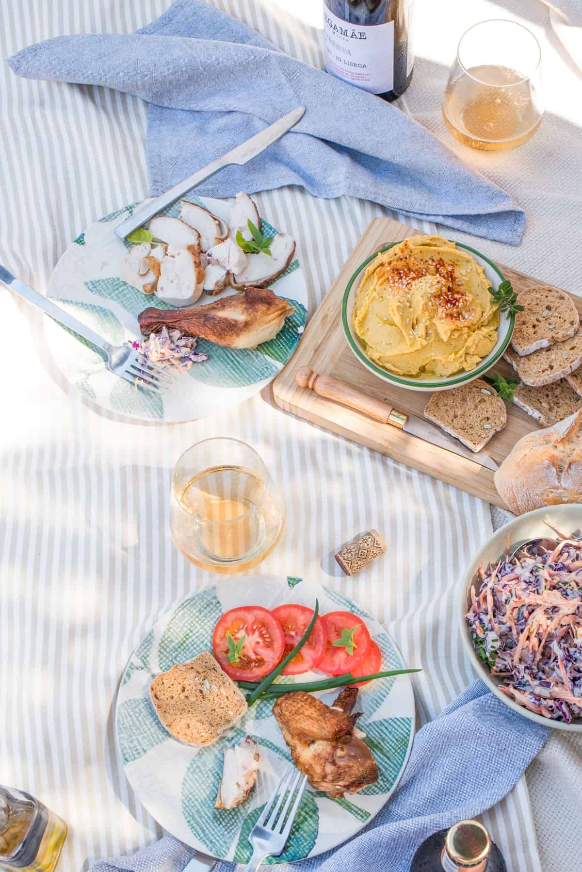 A picnic setup featuring two plates with chicken, mixed salads, and bread, a bowl of hummus with pita bread, two glasses of beverage, and utensils on a striped cloth.