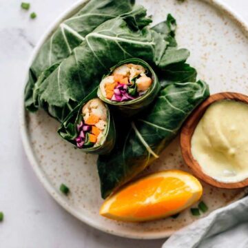 Collard wraps on a plate with a dipping sauce and an orange.