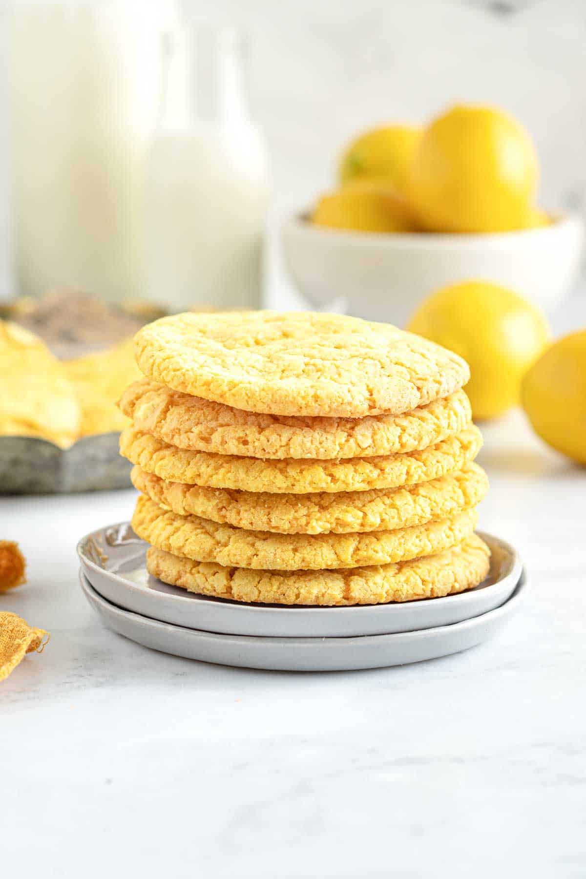 A stack of textured lemon crinkle cookies on a gray plate, with lemons and glass milk bottles in the background.