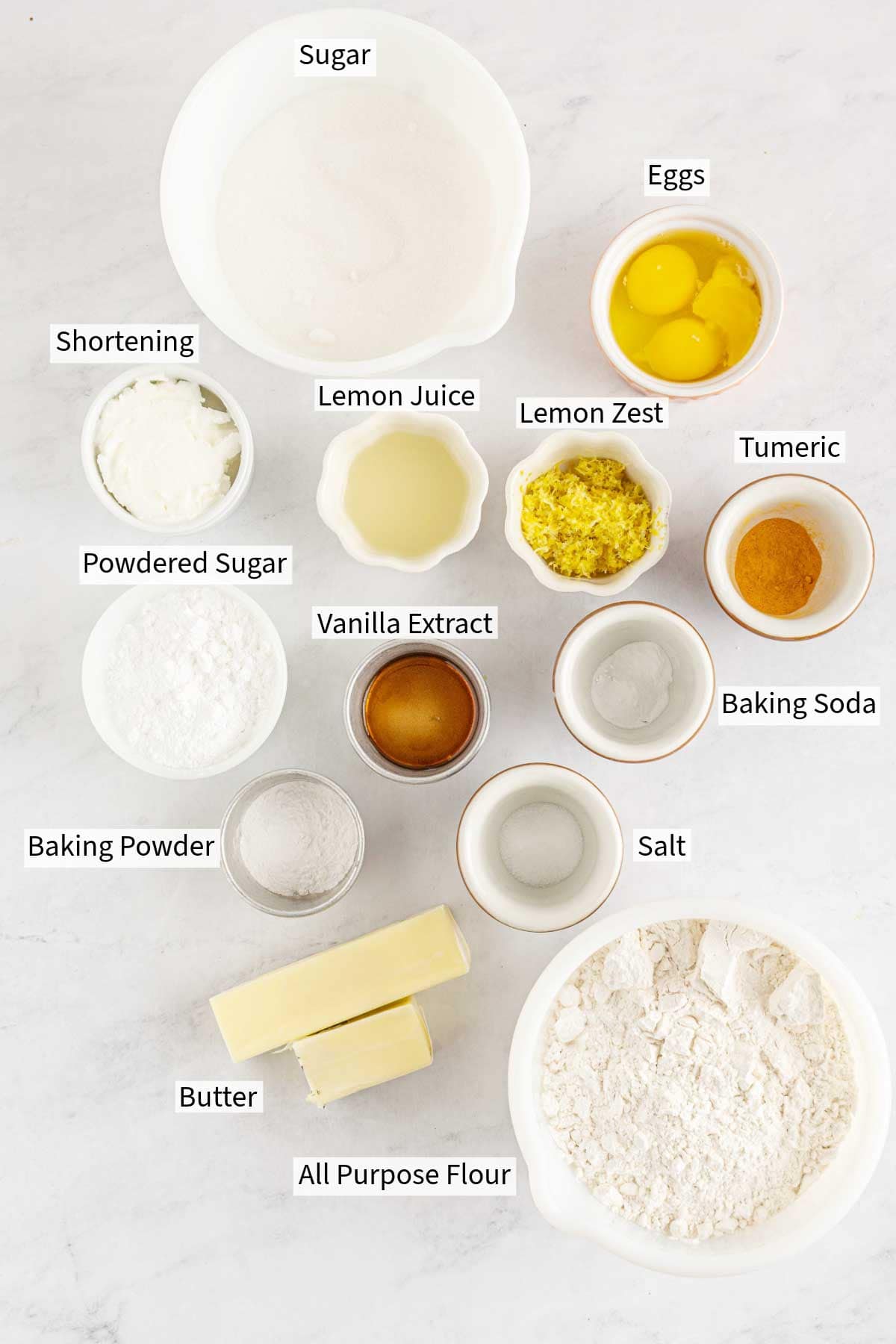 Overhead view of various baking ingredients for lemon crinkle cookies labeled in bowls on a white surface.
