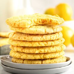 A stack of golden lemon sugar cookies on a small plate, with lemons blurred in the background.