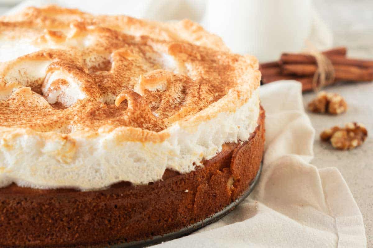 A pumpkin coffee cake topped with golden-brown meringue, surrounded with cinnamon sticks and walnuts on a light surface.