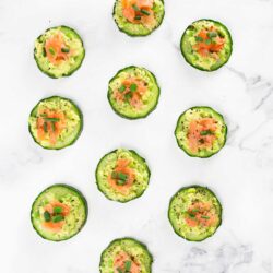 An array of sliced cucumber cups filled with guacamole and topped with smoked salmon, arranged neatly on a marble surface.