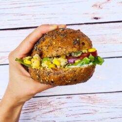 A hand holding a whole grain vegan tuna salad sandwich bun filled with chickpeas, lettuce, tomatoes, red onions, and a creamy sauce.