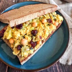 A vegan sandwich with chickpeas, avocado, and dried cranberries on whole grain bread, served on a blue plate with a rustic wooden background.