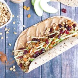 Open-faced sandwich with apple slices, granola, dried cranberries, and peanut butter on a whole wheat wrap, served on a wooden surface.