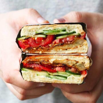 Hands holding a vegan panini sandwich with layers of tomatoes and cucumbers.