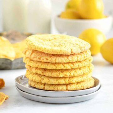 A stack of lemon cookies on a small plate, with lemons and a milk bottle in the background.