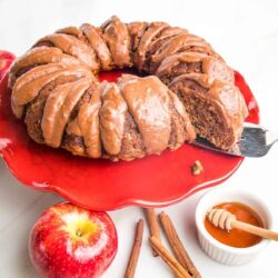 A frosted bundt cake on a red cake stand, surrounded by an apple, cinnamon sticks, and a small dish of honey with a dipper.