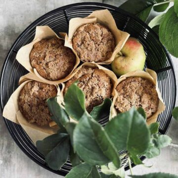 A black wire basket contains five muffins wrapped in brown paper and an apple. Green leaves partially cover the bottom of the basket.