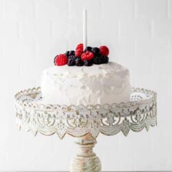 A white frosted sugar -free birthday cake topped with assorted berries and a single white candle, displayed on a decorative cake stand against a light background.