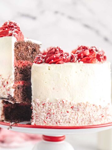 A chocolate and vanilla layered cake with white frosting and red rosettes is being sliced. The cake is decorated with crushed peppermint on the side. A bottle of milk is in the background.