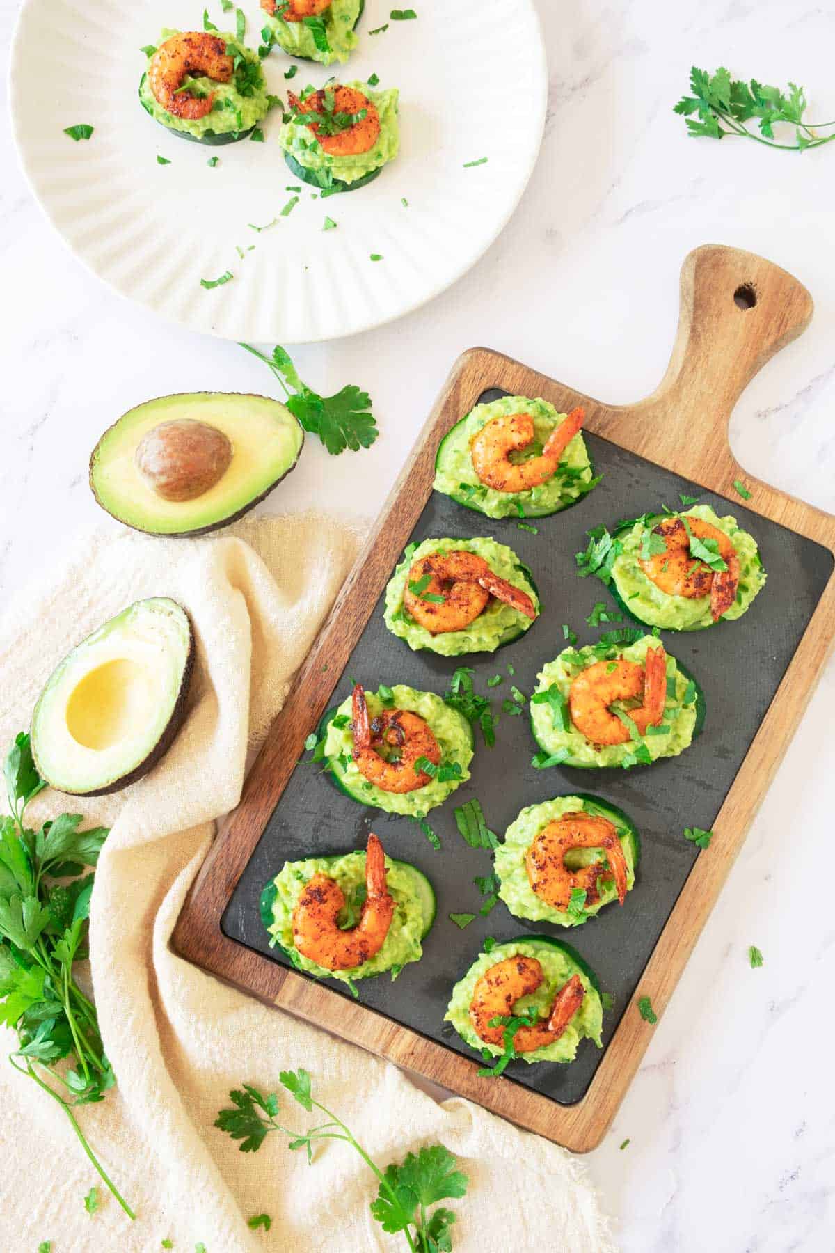 A wooden board with shrimp and avocado bite appetizers, surrounded by fresh avocados and herbs. 