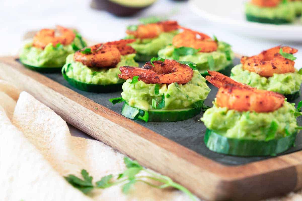 Slices of cucumber shrimp bites topped with guacamole and seasoned shrimp on a wooden board, garnished with chopped herbs.