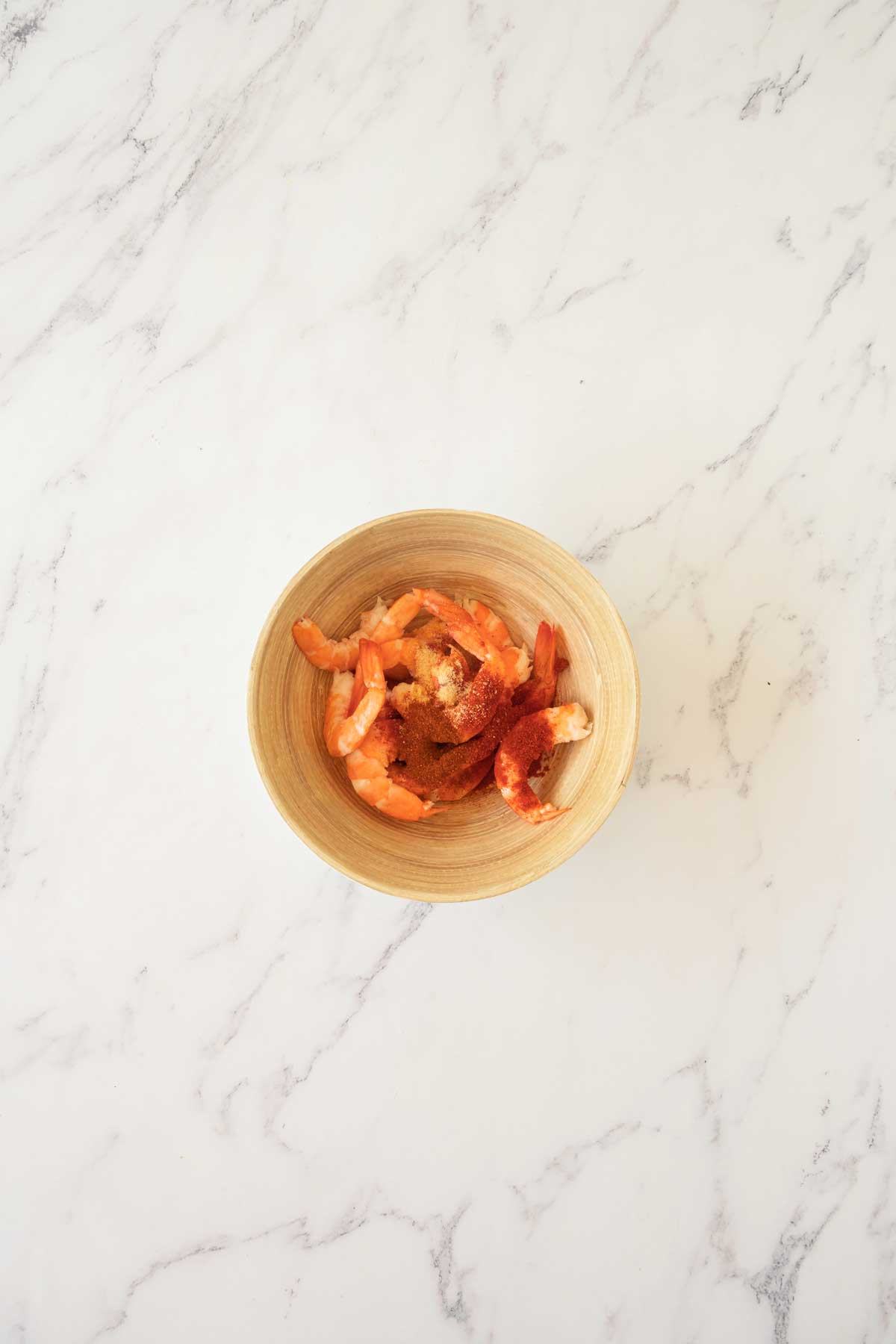 A wooden bowl filled with cooked shrimp seasoned with spices sits on a marble surface.