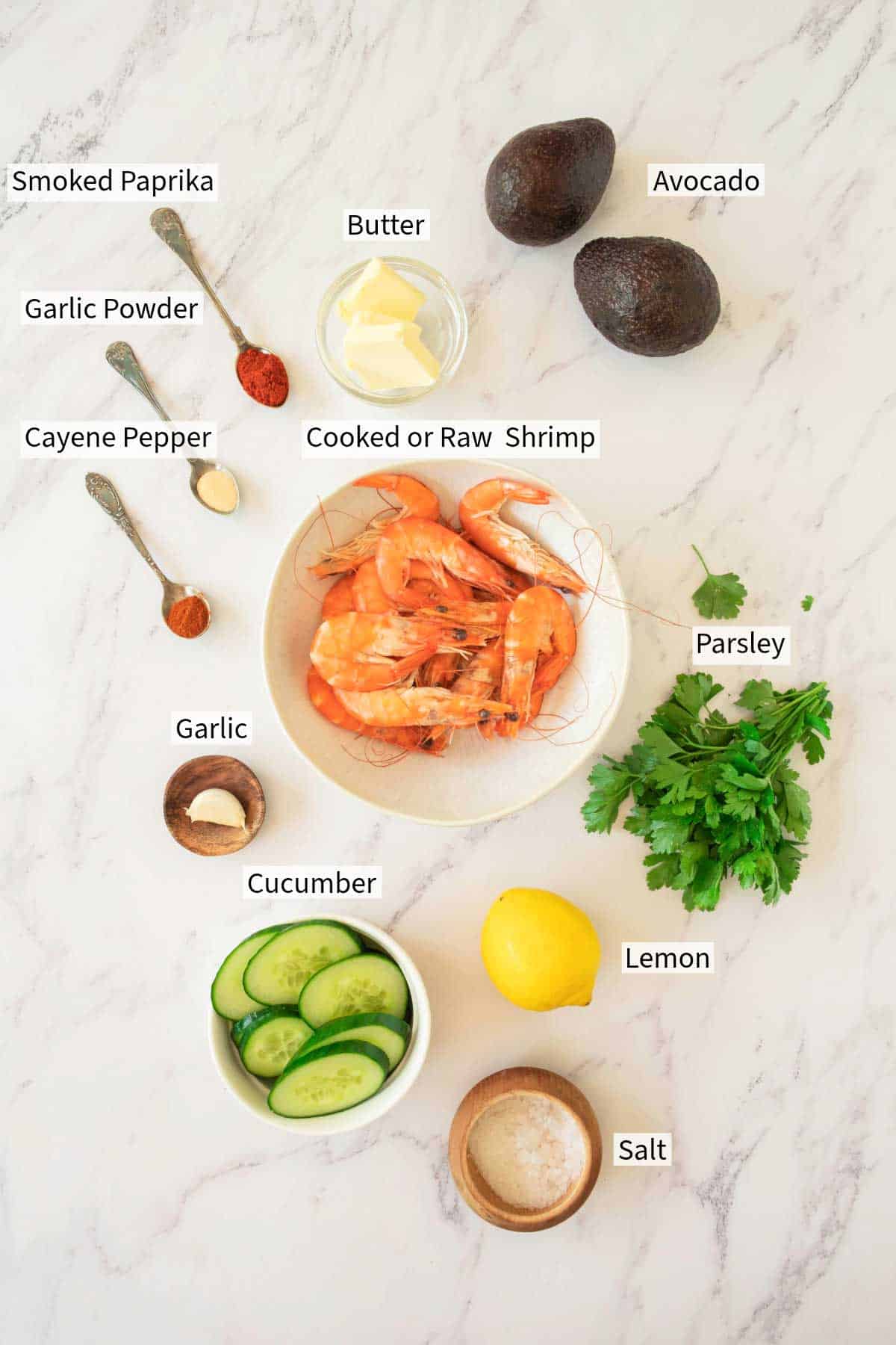 Ingredients laid out on a marble surface include smoked paprika, garlic powder, cayenne pepper, shrimp, butter, avocado, parsley, garlic, cucumber, lemon, and salt.