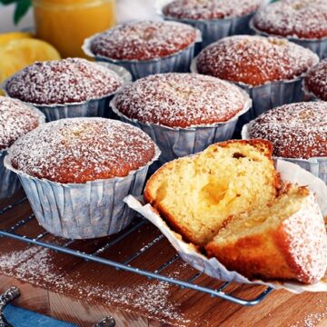 A batch of freshly baked muffins dusted with powdered sugar rests on a cooling rack, with one muffin partially unwrapped and cut in half to reveal its moist interior.