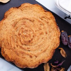 A golden brown date cake with a swirled crust, placed on a black tray alongside some dried figs or dates and scattered flower petals.