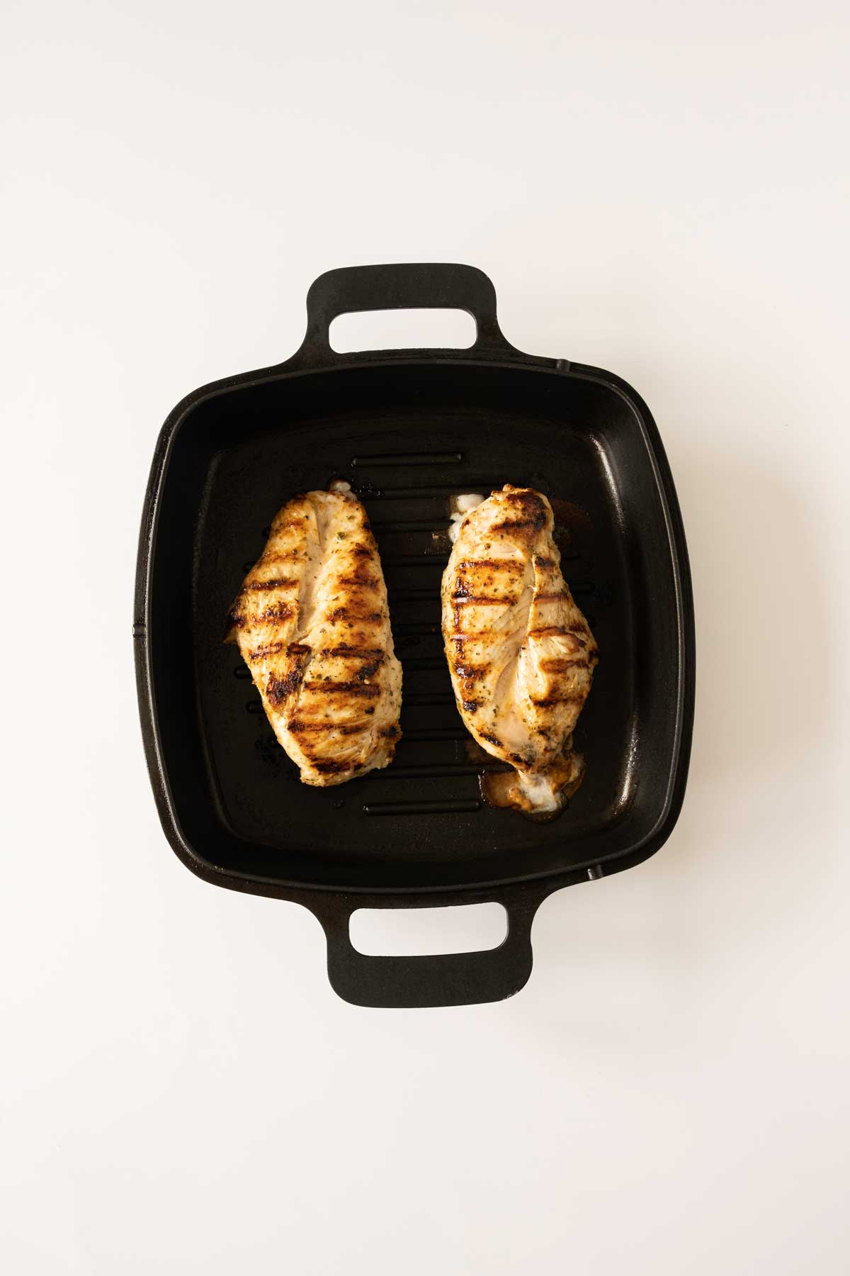 Two grilled chicken breasts with grill marks are placed side by side in a black grill pan on a white background.