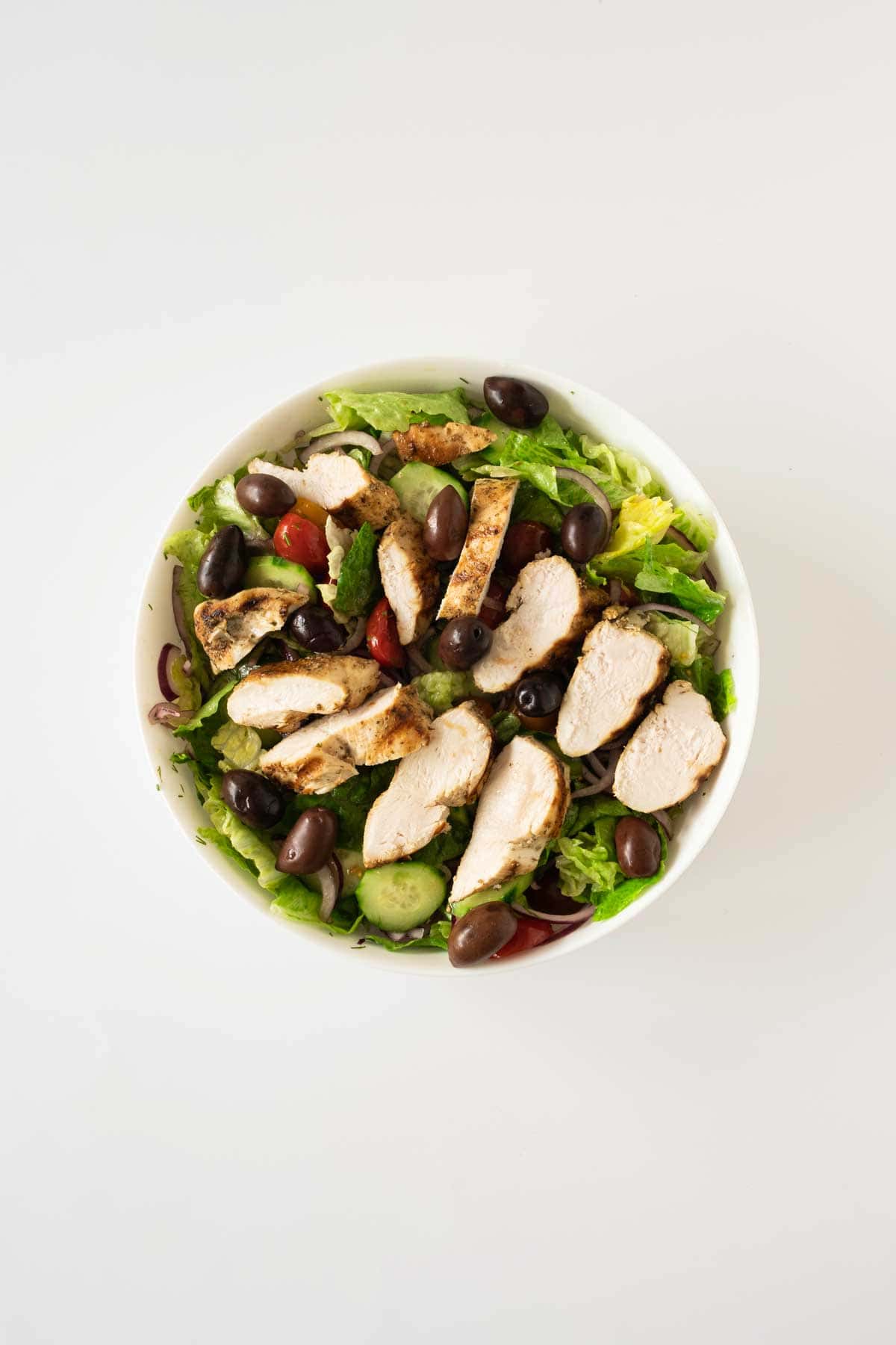 A bowl of mixed salad with grilled chicken strips, black olives, cherry tomatoes, and cucumber slices on a white background.