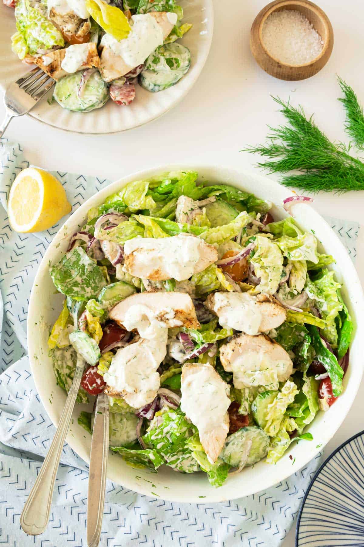 A bowl of Greek Chicken salad with chicken slices, lettuce, tomatoes, and dressing, accompanied by a squeezed lemon, herbs, and a small dish of salt. 