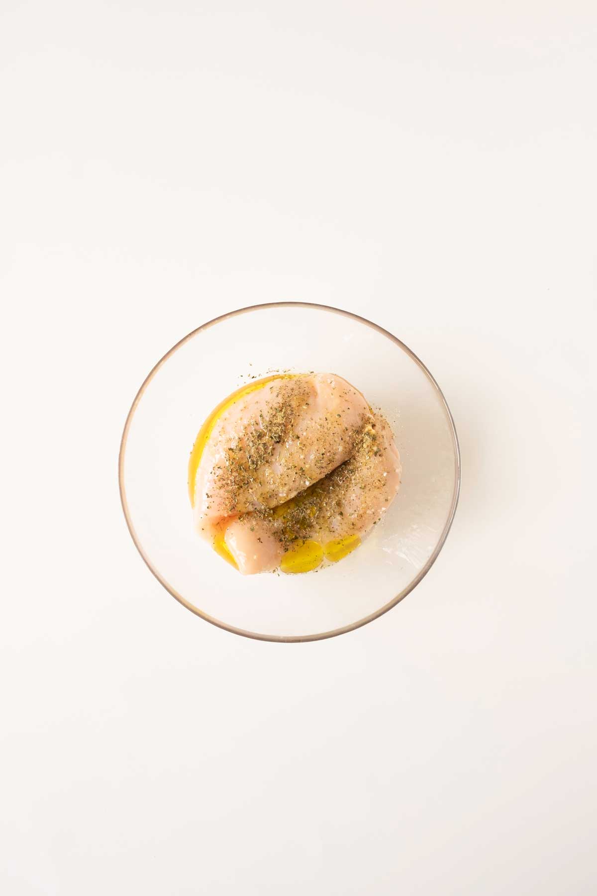 Raw seasoned chicken breast in a glass bowl on a white background.