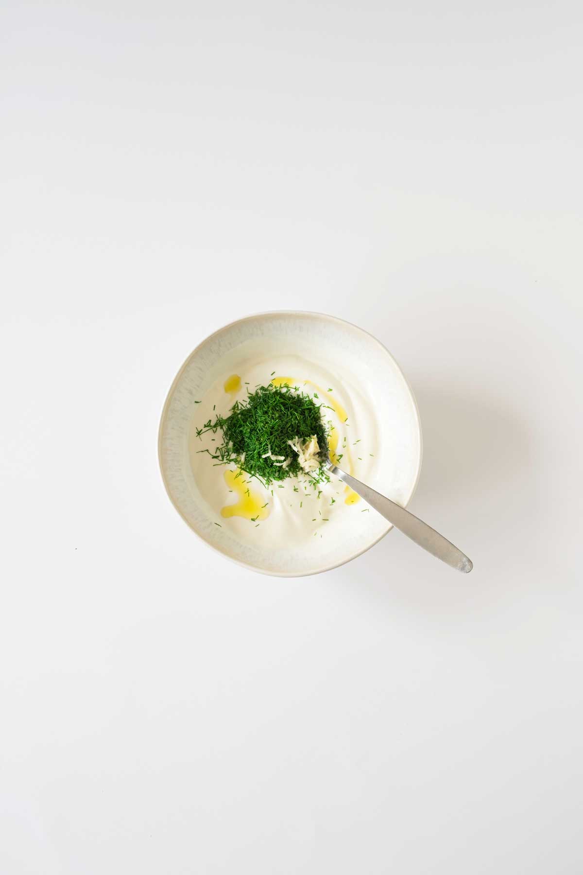 A bowl of yogurt with herbs on top and a spoon resting inside.