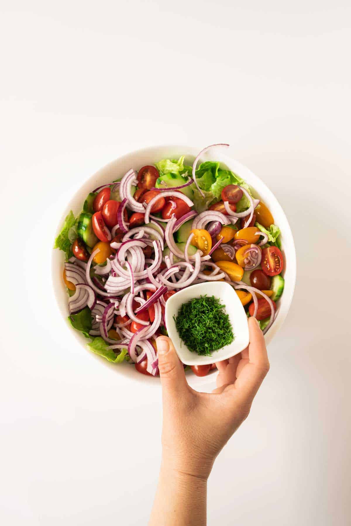 A hand holding a small white bowl of chopped herbs over a salad with lettuce, cherry tomatoes, red onions, and cucumber slices.