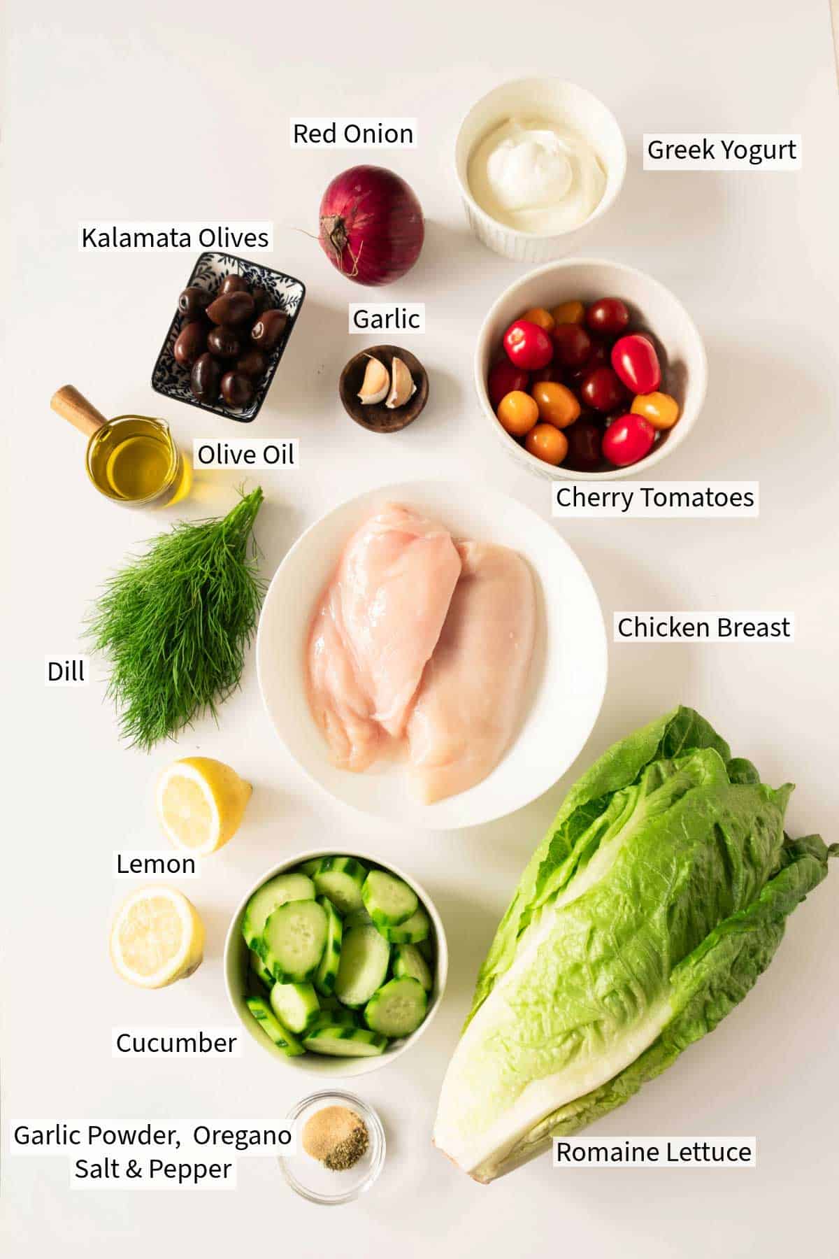 Ingredients for Greek Chicken Salad with Tzatziki Dressing on a white surface, including chicken breast, romaine lettuce, cucumber, cherry tomatoes, red onion, garlic, Greek yogurt, kalamata olives, and seasonings.