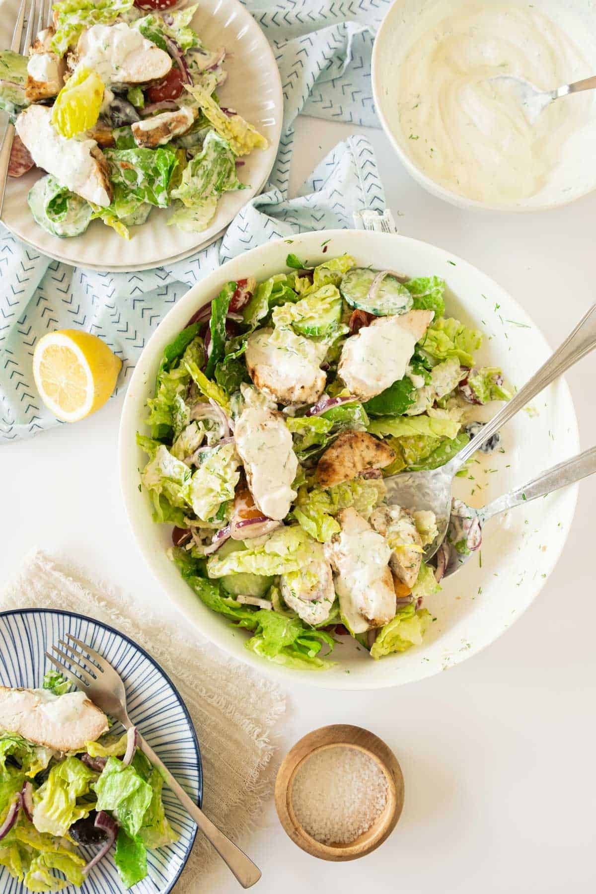 A table featuring three plates of Greek Chicken Salad, a lemon half, a small wooden bowl of salt, and a bowl of creamy dressing with a spoon.