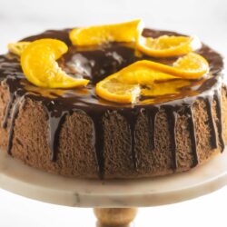 A chocolate cake with a glossy chocolate glaze, garnished with orange slices, sits on a marble cake stand.