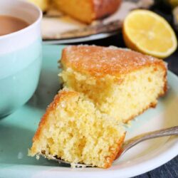 A slice of lemon cake on a plate with a fork beside a cup of tea and a sliced lemon in the background.