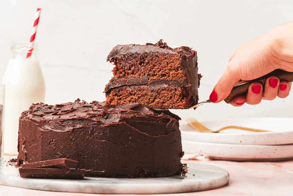 A hand lifts a slice of Matilda chocolate cake with a cake server. The cake is frosted with chocolate icing.