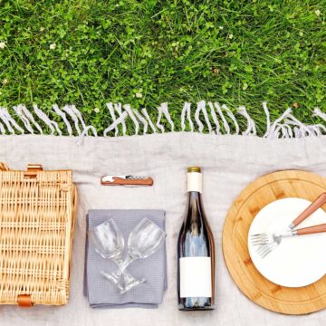 A picnic setup on a grassy area with a woven basket, two wine glasses, a bottle of wine, a corkscrew, a cutting board with cutlery, and a gray cloth.