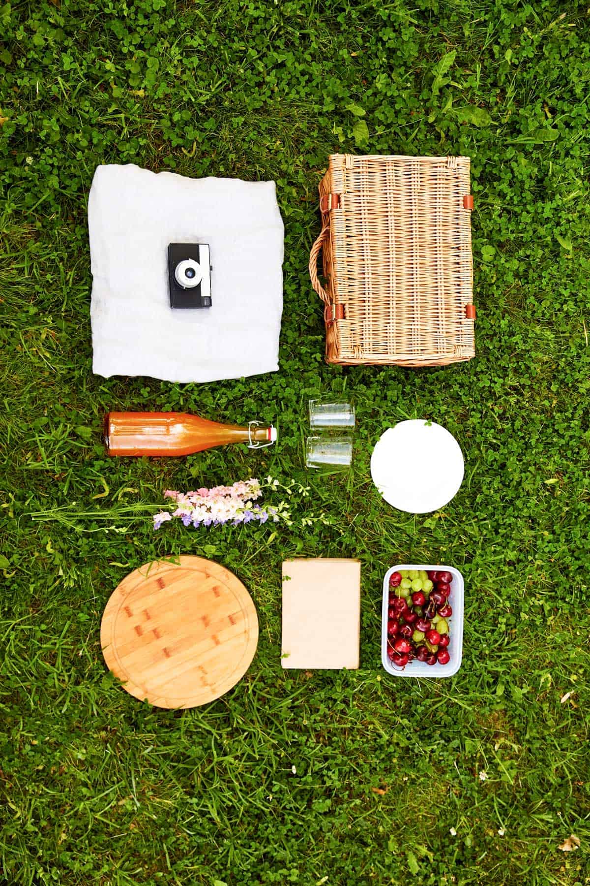 A picnic setup on grass with a wicker basket, white cloth, two glasses, a wooden tray, a bottle of drink, a container of grapes and cherries, purple flowers, and a small square of cheese.