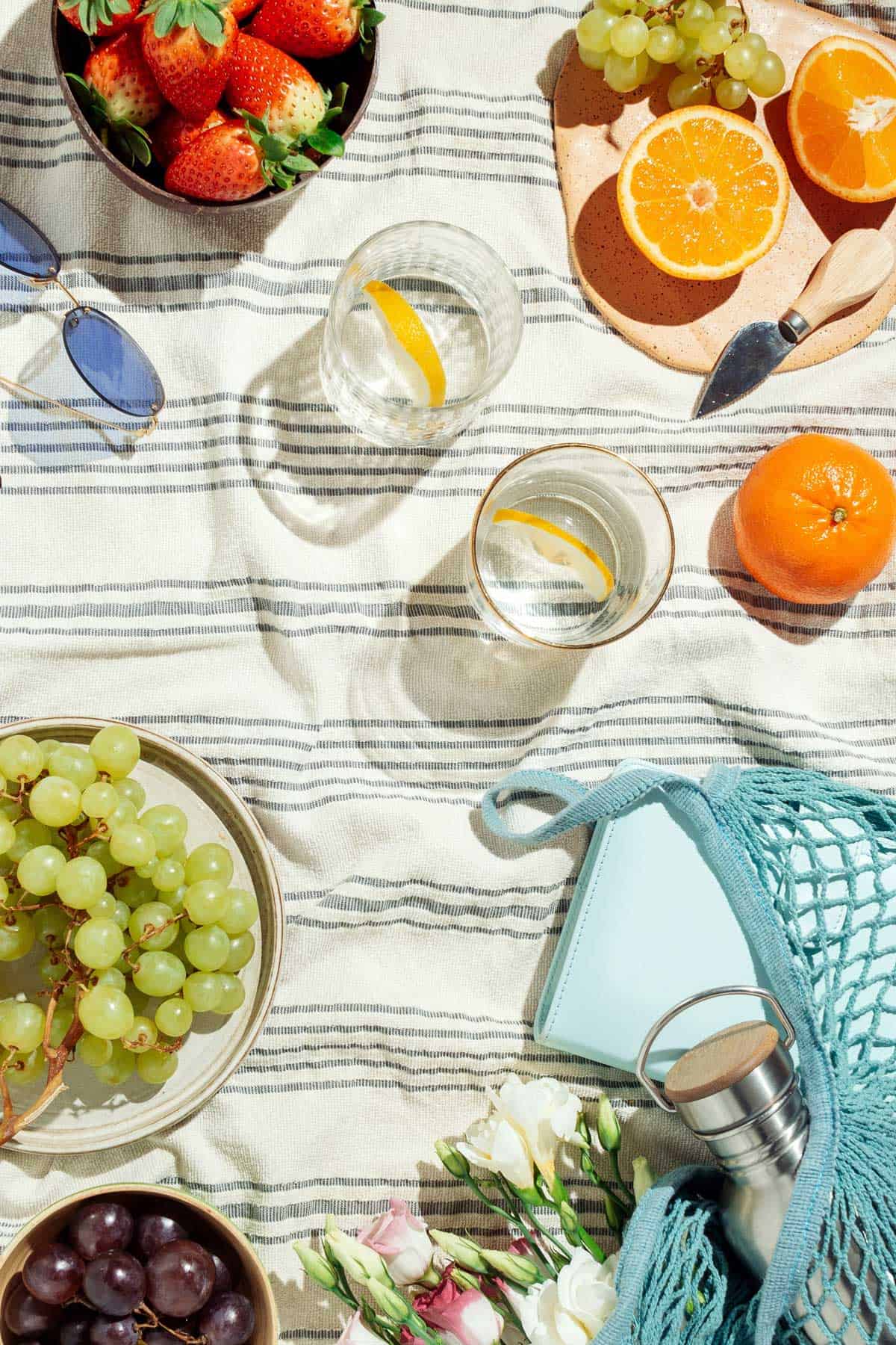 A picnic blanket with bowls of strawberries, grapes, oranges, sunglasses, two glasses of lemon water, a blue bag, flowers, and a journal.