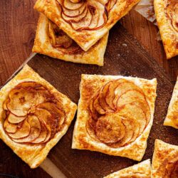 Squares of puff pastry topped with thinly sliced apples and a sprinkle of cinnamon are displayed on a wooden surface.