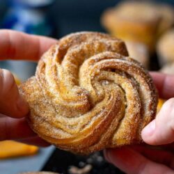 Close-up of hands holding a swirled pastry covered in sugar, with some additional pastries visible in the background.