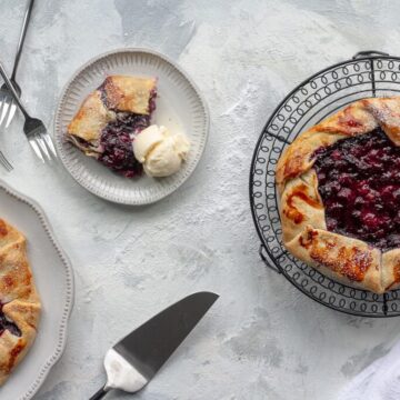 A baked galette with a berry filling on a cooling rack, a serving on a plate with a scoop of ice cream, and kitchen utensils on a textured surface.