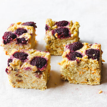 Four square pieces of dessert with a crumbly texture and topped with dark red fruit are placed on a white surface.