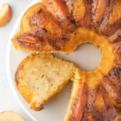 A round peach upside-down cake with one slice removed, showing the interior. The cake has a golden-brown top with peach slices arranged in a circular pattern.