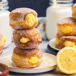 A stack of three sugar-coated donuts filled with yellow custard is placed on a plate. A jar of milk and a slice of lemon are visible in the background.