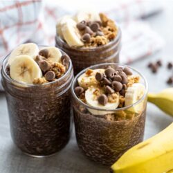 Three glass jars filled with chocolate chia pudding, topped with banana slices, granola, and chocolate chips, with bananas and a checkered cloth in the background.