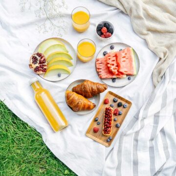 A picnic setup on a white blanket with sliced melon, watermelon, pomegranate seeds, croissants, juice, and bowls of blackberries and raspberries.