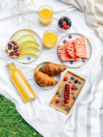 A picnic setup on a white blanket with sliced melon, watermelon, pomegranate seeds, croissants, juice, and bowls of blackberries and raspberries.