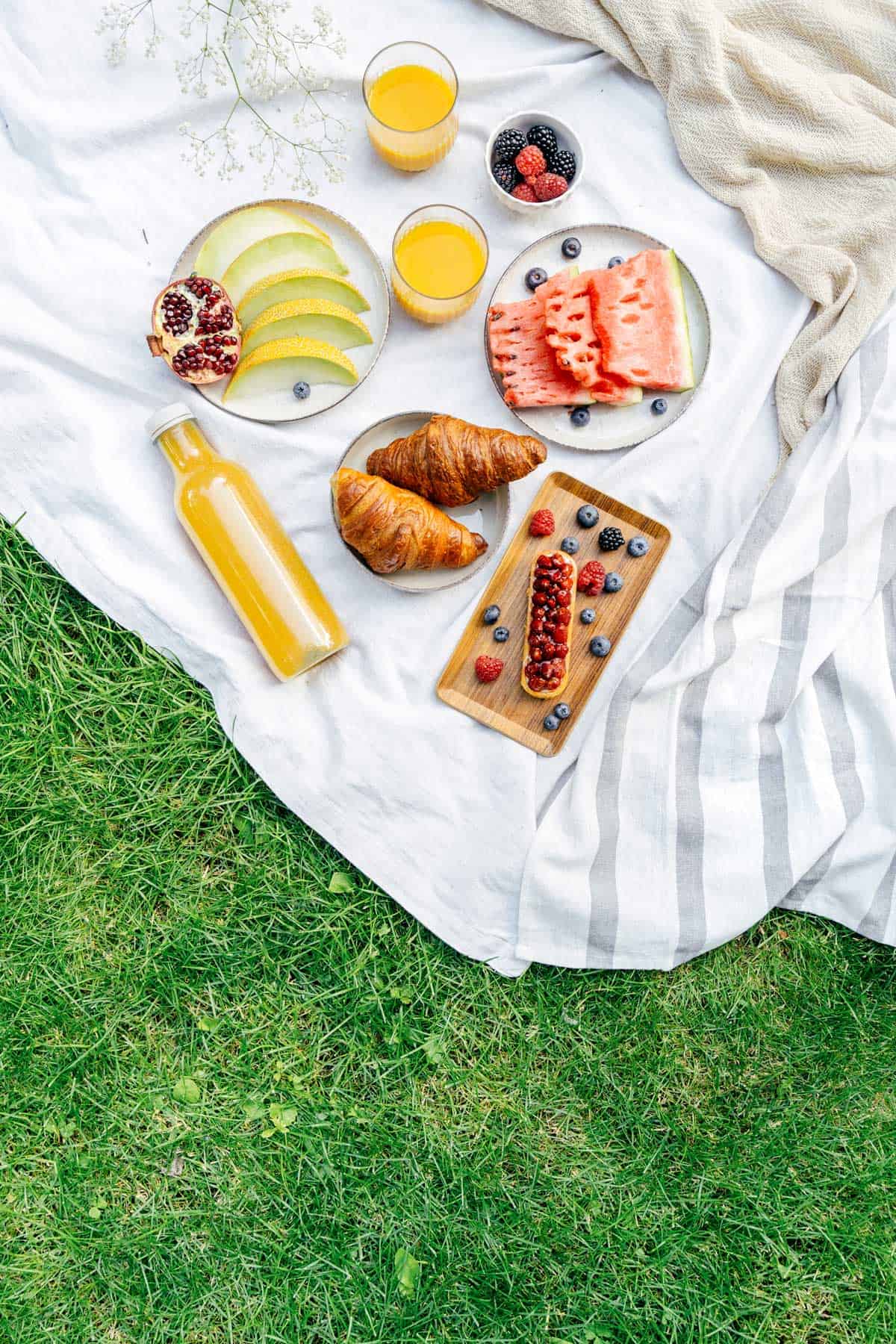 A breakfast picnic setup on a blanket with croissants, watermelon, melon, berries, pomegranate, juice, and two glasses of juice on grass.